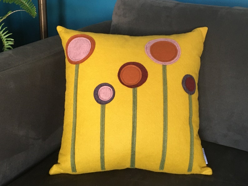 K&ouml;nnte beinhalten: Ein gelbes Kissen mit einem Design aus f&uuml;nf stilisierten Blumen in den Farben Rosa, Orange, Braun und Lila. Die Blumen sind gr&uuml;n umrandet.