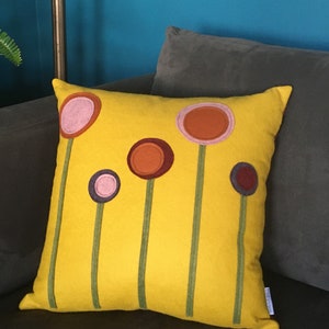 K&ouml;nnte beinhalten: Ein gelbes Kissen mit f&uuml;nf bunten Filzkreisen, die wie Blumen aussehen, auf der Vorderseite. Die Kreise sind in den Farben Rosa, Orange, Braun, Lila und Rotbraun.