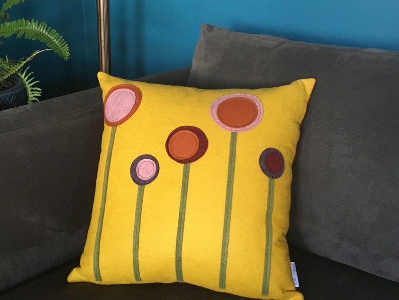 K&ouml;nnte beinhalten: Ein gelbes Kissen mit f&uuml;nf bunten Filzkreisen, die wie Blumen aussehen, auf der Vorderseite. Die Kreise sind in den Farben Rosa, Orange, Braun, Lila und Rotbraun.