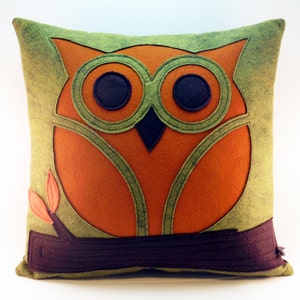 May include: A green and orange felt pillow with an owl design. The owl is sitting on a brown branch with two small leaves.