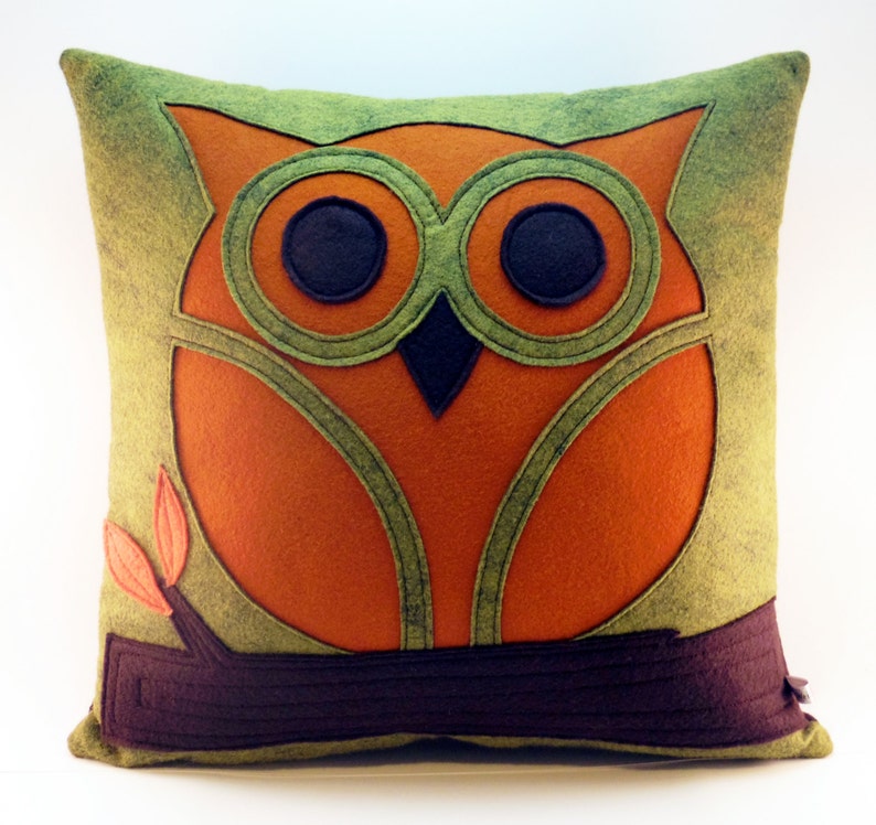 May include: A green and orange felt pillow with an owl design. The owl is sitting on a brown branch with two small leaves.