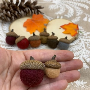 May include: Handmade felt acorns in various colors, including burgundy, orange, and gray, with natural acorn caps. The acorns are displayed with wooden slices and fall foliage, evoking an autumn theme.