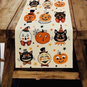 May include: A cream-colored table runner featuring a repeating pattern of vintage-style Halloween illustrations, including black cats with bow ties, orange jack-o'-lanterns, ghosts in party hats, and moon faces, all accented with small orange and black stars.