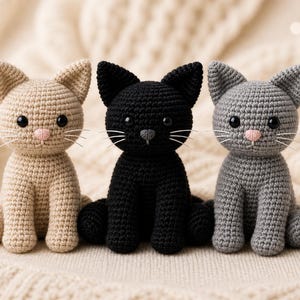 May include: Three crocheted cat figurines in beige, black, and gray. Each cat has large black eyes, pink noses, and white whiskers. The cats are sitting upright on a soft, textured surface, showcasing the handmade details.