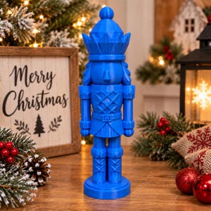 May include: A bright blue nutcracker figurine featuring a geometric, faceted design. The nutcracker wears a tall, pointed hat with a pom-pom top, a jacket with a cross-hatched pattern, a belt, and tall boots, all standing on a circular base.