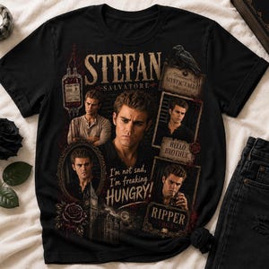 Puede incluir: Camiseta negra con un diseño que presenta a Stefan Salvatore. El diseño incluye retratos, las palabras "STEFAN SALVATORE", "MYSTIC FALLS", "HELLO BROTHER" y "RIPPER". También es visible la frase "I'm not sad, I'm freaking HUNGRY!".