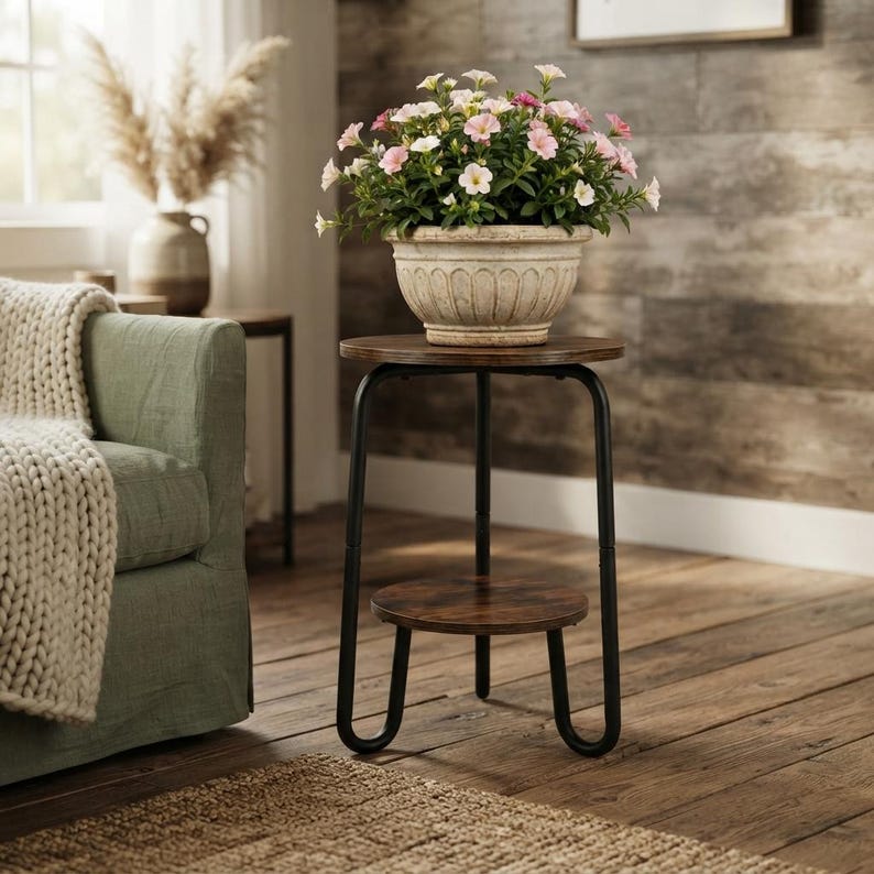 Pu&ograve; includere: Un tavolino rotondo con piano in legno scuro e ripiano inferiore, sostenuto da una struttura in metallo nero con tre gambe a U. Un vaso in ceramica scanalata color crema contiene fiori di petunia rosa e bianchi sulla superficie.