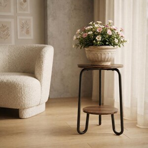 Pu&ograve; includere: Un portapiante rotondo in legno con finitura marrone scuro e telaio in metallo nero con ripiano inferiore. Un vaso in ceramica beige scanalato poggia sulla superficie superiore, contenente piccoli fiori rosa chiaro e bianchi con foglie verdi.
