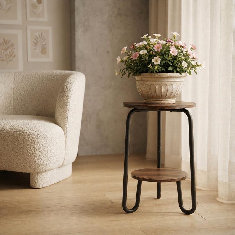 Pu&ograve; includere: Un portapiante rotondo in legno con finitura marrone scuro e telaio in metallo nero con ripiano inferiore. Un vaso in ceramica beige scanalato poggia sulla superficie superiore, contenente piccoli fiori rosa chiaro e bianchi con foglie verdi.