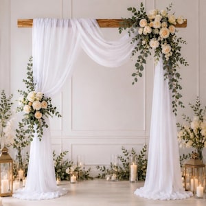 Può includere: Un arco nuziale con drappeggi in tessuto bianco e composizioni floreali. L'arco è realizzato con una trave di legno ed è decorato con rose bianche e verde. Candele e lanterne sono disposte attorno alla base, creando un'atmosfera romantica.