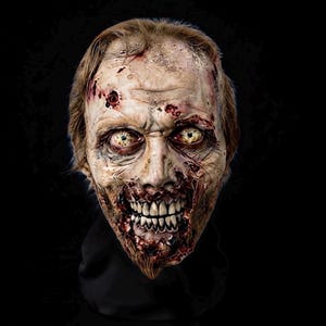 May include: A realistic zombie mask featuring pale, decaying skin with visible wounds, sores, and bloodstains. The mask has sunken, yellowed eyes, a wide, toothy grin with exposed gums, and sparse, light brown hair on the top of the head.