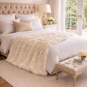 May include: A bedroom with a tufted headboard, white bedding, and a cream-coloured faux fur throw. A matching bench with gold legs is at the bed's foot. A lamp and flowers complete the elegant decor, creating a serene atmosphere.