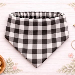 May include: A black and white checkered dog bandana with an elastic neckline. The bandana is surrounded by dog treats, a wooden bowl, and decorative floral elements. A pair of gold scissors and a string are also present.