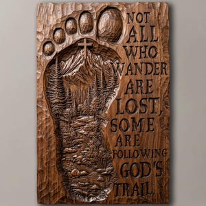 Puede incluir: Una placa de pared rectangular de madera marrón con una huella tallada. Dentro de la huella hay una escena en relieve de una montaña, un bosque y una cruz. El texto dice: NOT ALL WHO WANDER ARE LOST, SOME ARE FOLLOWING GOD'S TRAIL.