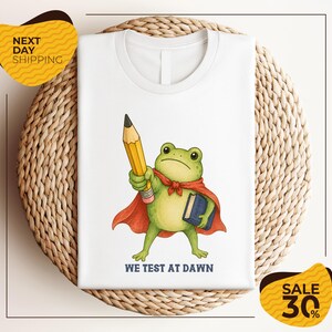 May include: A white t-shirt featuring a graphic of a green frog wearing a red cape, holding a yellow pencil in one hand and a blue book in the other. Below the frog, the text reads WE TEST AT DAWN in dark blue capital letters.