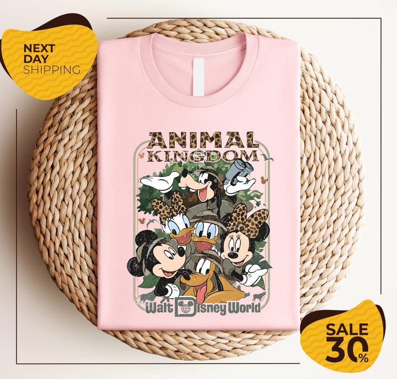 A pink t-shirt with a Disney-themed graphic design, featuring characters from the Disney movie "Animal Kingdom" and the text "Walt Disney World". The t-shirt is displayed on a woven basket, and there are two yellow sale signs indicating a 30% discount.