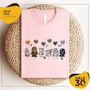 A pink t-shirt with a graphic design featuring various Star Wars characters, including Chewbacca, Stormtrooper, Darth Vader, and Baby Yoda, along with other Star Wars-themed elements.