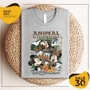 A gray t-shirt with a graphic design featuring Disney characters Mickey Mouse, Minnie Mouse, Donald Duck, and Goofy, along with the text "Animal Kingdom" and " Walt Disney World". The t-shirt is displayed on a woven placemat, and there are yellow and black sale signs indicating a 30% discount.