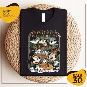 A black t-shirt with a graphic design featuring various Disney characters, including Mickey Mouse, Minnie Mouse, Donald Duck, Goofy, and Pluto, arranged in a circular frame.