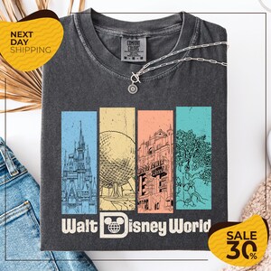 A gray t-shirt with a graphic design featuring various Disney landmarks, including the Eiffel Tower, the Walt Disney World resort, and a tree. The t-shirt is displayed on a white background, and there are jeans and a sweater visible in the background.