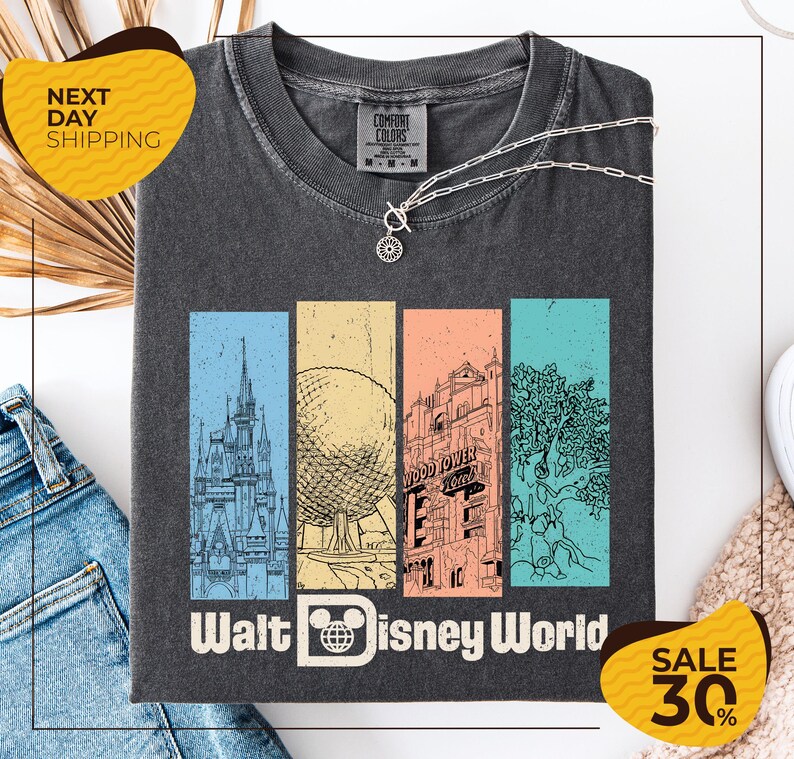 A gray t-shirt with a graphic design featuring various Disney landmarks, including the Eiffel Tower, the Walt Disney World resort, and a tree. The t-shirt is displayed on a white background, and there are jeans and a sweater visible in the background.