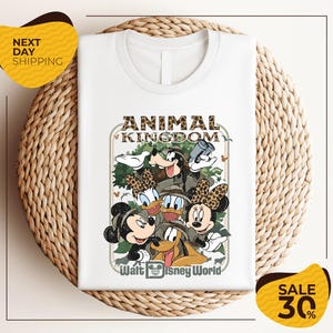 A white t-shirt with a Disney-themed graphic design featuring Minnie Mouse, Donald Duck, and Goofy, along with the text "Animal Kingdom" and " Walt Disney World". The t-shirt is displayed on a woven placemat, and there are yellow sale signs indicating a 30% discount.