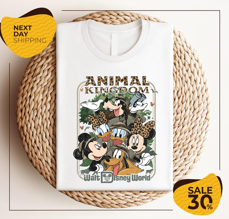 A white t-shirt with a Disney-themed graphic design featuring Minnie Mouse, Donald Duck, and Goofy, along with the text "Animal Kingdom" and " Walt Disney World". The t-shirt is displayed on a woven placemat, and there are yellow sale signs indicating a 30% discount.