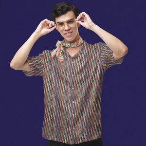 May include: A short-sleeved button-down shirt featuring a vertical rectangular pattern in shades of blue, orange, yellow, and brown. The shirt has a notched collar and is paired with a patterned neck scarf and clear-framed aviator-style glasses.