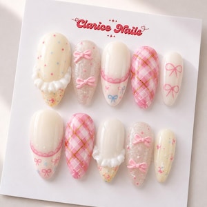 May include: A set of ten almond-shaped press-on nails on a white display card labeled Clarice Nails. The designs feature cream, pink, and glitter finishes with 3D white ruffles, pink bows, gold grid lines, and small colorful polka dot patterns.
