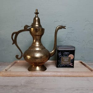 Peut inclure: Une théière en laiton vintage avec des gravures florales complexes, un long bec et une poignée décorative. À côté, une boîte de thé noire avec le texte "THE DE NOEL CANNELLE". Les articles sont sur un plateau en bois.