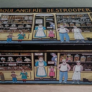Peut inclure: Une boîte de rangement rectangulaire en métal ornée d'une illustration noire et jaune d'une devanture de boulangerie intitulée BOULANGERIE DESTROOPER. Le décor représente des boulangers, des clients et des étagères remplies de pâtisseries.