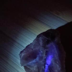 May include: A rough-cut, dark purple fluorite crystal with a bright blue, glowing vein. The crystal is set against a wooden surface with a subtle, diagonal grain pattern. The lighting highlights the crystal's texture.