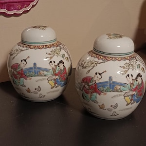 Peut inclure: Deux pots à gingembre en céramique blanche avec couvercles assortis, ornés d'illustrations peintes à la main représentant des personnages en vêtements traditionnels interagissant avec de petits oiseaux dans un jardin, avec des motifs floraux.