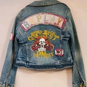 Può includere: Giacca di jeans blu chiaro con un aspetto vissuto. La schiena presenta una grafica con un teschio, chitarre e la scritta "Hard Rock Cafe Detroit 1984". Sono presenti anche toppe con "REPLAY" e "MG".