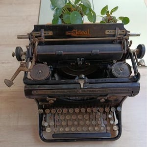 Può includere: Un'antica macchina da scrivere nera con la parola "Ideal" visibile. La macchina da scrivere ha una tastiera completa con tasti rotondi e una leva di ritorno del carrello. Una piccola pianta verde è posta sopra.