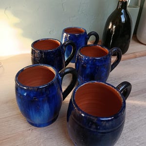 Könnte beinhalten: Ein Set aus vier fassförmigen Keramikbechern mit einer glänzenden tiefblauen Glasur auf der Außenseite und einem natürlichen Terrakotta-Finish auf der Innenseite. Jeder Becher verfügt über einen stabilen, geschwungenen Henkel und einen runden Körper.