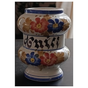 May include: A ceramic vase with a bulbous shape, featuring hand-painted floral designs in red, blue, and gold against a cream background. The vase has a blue rim and the word "Kniff" is written in black script.