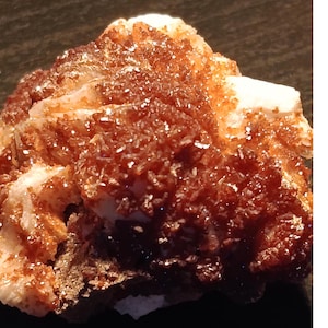 Puede incluir: Primer plano de un espécimen mineral que presenta un grupo de cristales de color marrón rojizo. Los cristales están densamente empaquetados y brillan, contrastando con la roca base más clara, blanquecina. La textura general es áspera y desigual.