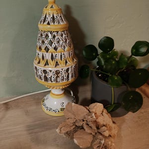 Puede incluir: Una lámpara de mesa de cerámica con una pantalla cónica alta y calada, decorada con motivos florales pintados a mano en amarillo y azul. La lámpara tiene una base circular a juego y un cable blanco. Junto a ella hay un racimo de cristales.