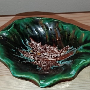 Puede incluir: Un cuenco de cerámica con forma de hoja y acabado brillante. El cuenco presenta un exterior verde oscuro y un interior con estampado marrón y blanco. El cuenco está sobre una superficie de madera clara.