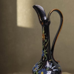 Op de afbeelding: Een donkerblauwe keramische kan met een slanke hals en een gebogen handvat. De kan is versierd met handgeschilderde bloemmotieven in blauw, geel en oranje. Het handvat heeft een bruine rand. Deze decoratieve kan is een uniek stuk.