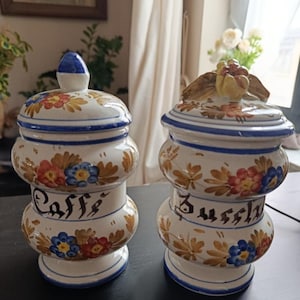 Peut inclure: Deux pots en céramique avec couvercles. Les pots sont blancs avec des bordures bleues et présentent des motifs floraux rouges, jaunes et bleus. L'un des pots porte l'inscription "Caffe" et l'autre "Biscotti". Les couvercles ont des ornements décoratifs.