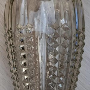 Peut inclure: Un vase en verre transparent avec une surface texturée. Le vase présente un motif de losanges sur la moitié inférieure et des lignes verticales sur la moitié supérieure. Le vase est vide et reflète la lumière.