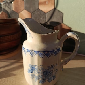 May include: A cream-colored ceramic pitcher featuring a blue floral pattern and a geometric border design. The pitcher has a curved handle, a pouring spout, and a visible crackled glaze finish across its surface, showcasing a classic aesthetic.
