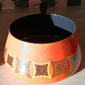 Pode incluir: Uma tigela vintage de esmalte laranja e vermelho com interior preto e um padrão floral decorativo no exterior. A tigela tem formato arredondado e uma única alça, perfeita para servir ou expor.