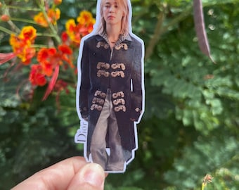 May include: A die-cut sticker featuring a person with light hair wearing a dark, long-sleeved jacket with ornate gold-colored embroidery and light-colored trousers. The word Dior is printed vertically in white along the left side of the sticker.