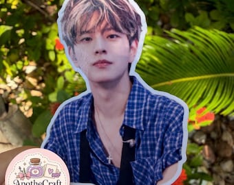 May include: A die-cut sticker featuring a portrait of a person with brown hair, wearing a blue and white plaid shirt and a silver cross necklace. The sticker has a white border and is held against a background of green foliage and red flowers.