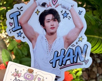 May include: A die-cut sticker featuring a person with dark hair wearing a white sleeveless top, arms raised above their head, surrounded by blue text reading I and HAN, plus star and heart doodles. A small rectangular business card reads ApotheCraft Studio.