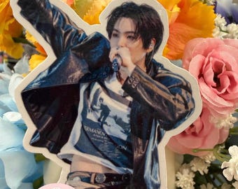 May include: A die-cut sticker featuring a person with dark hair performing into a microphone while wearing a black leather jacket over a white graphic t-shirt. The person is captured in a dynamic pose with one arm raised, showcasing a detailed print.