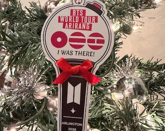 May include: A flat, light-colored ornament shaped like a lightstick, featuring the text BTS WORLD TOUR ARIRANG, I WAS THERE!, ARLINGTON 2026, and the BTS logo. It is decorated with a small red ribbon bow and hangs from a red and white string.